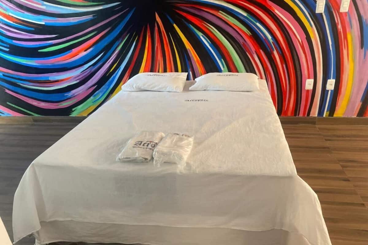 A neatly made bed with white bedding and pillows, a white table with a tray, and a colorful abstract wall art in the background.