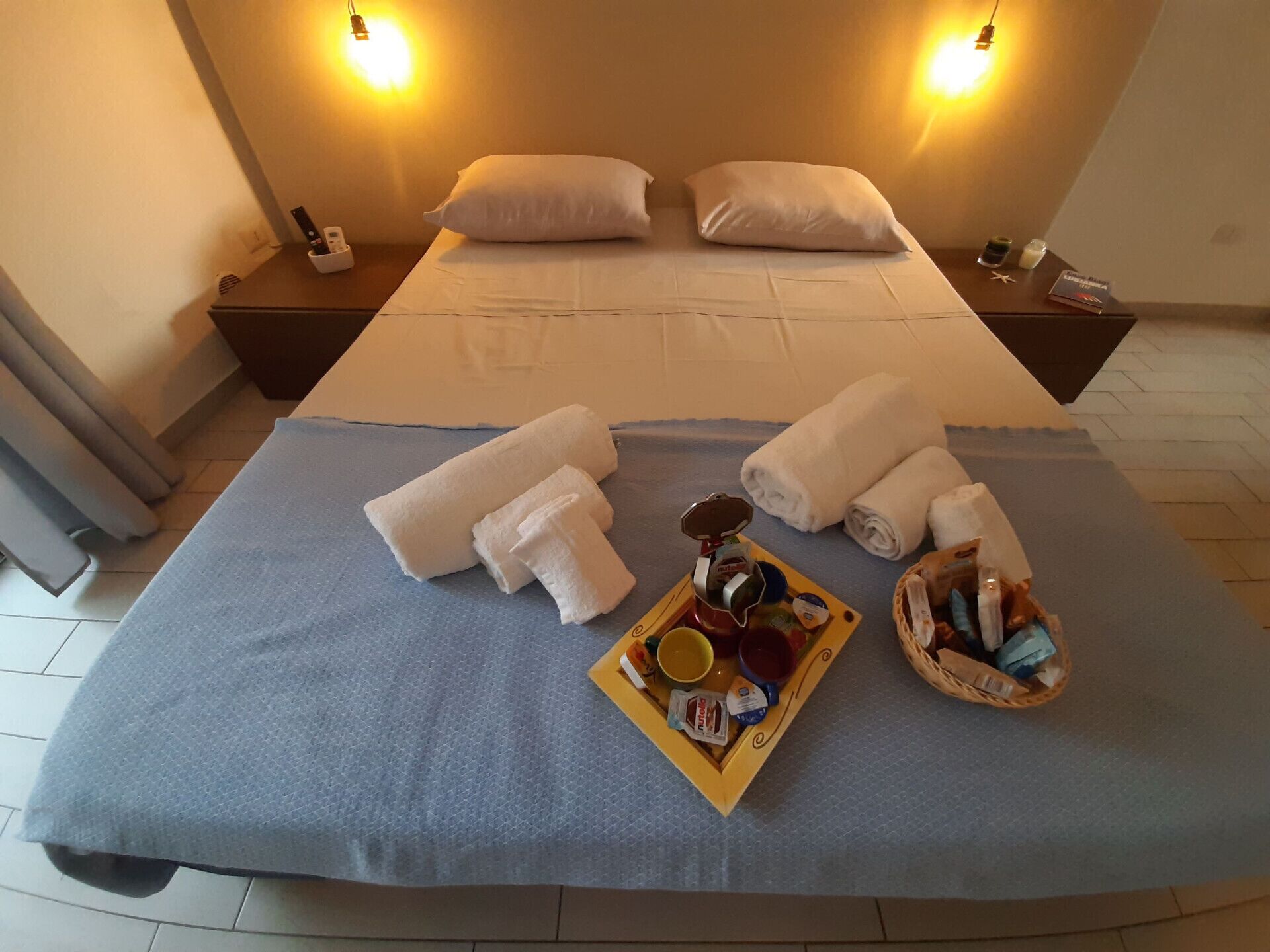A neatly made bed with white pillows and a blue bedspread, two bedside tables with lamps, and a tray of breakfast items on the bed.