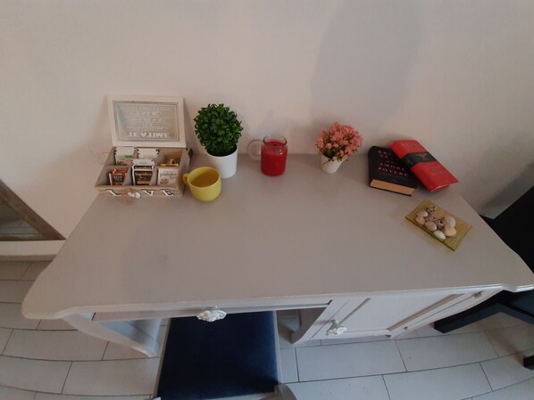 A small, white desk with a laptop, a potted plant, a red candle, a yellow mug, a framed picture, a small vase with pink flowers, a book, and a decorative plate with seashells.