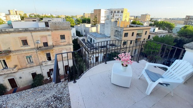 Een balkon met witte stoelen en een bloempot, met uitzicht op een stadsgezicht met verschillende gebouwen.