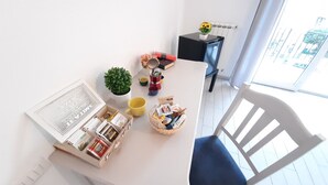 Een lichte kamer met een witte tafel, een kleine plant in een pot, een mand met lekkernijen en een raam met uitzicht op gebouwen.