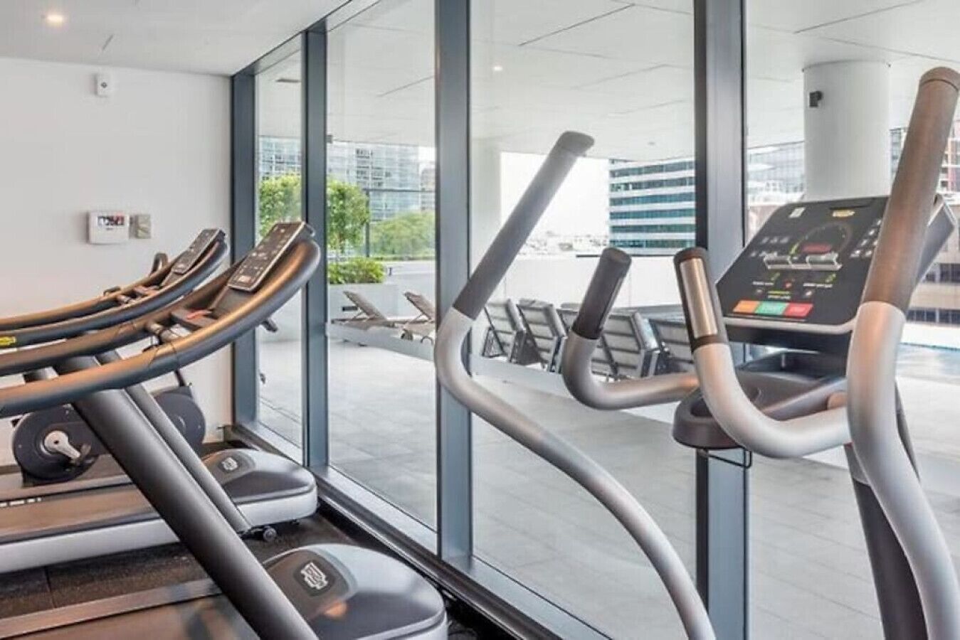 Un gimnasio moderno con cintas de correr y equipo de ejercicio, ventanas amplias y vista a la ciudad.