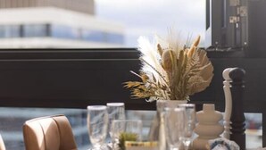 Une table à manger dressée avec des verres, des assiettes et un vase contenant des plantes séchées, donnant sur un bâtiment moderne.