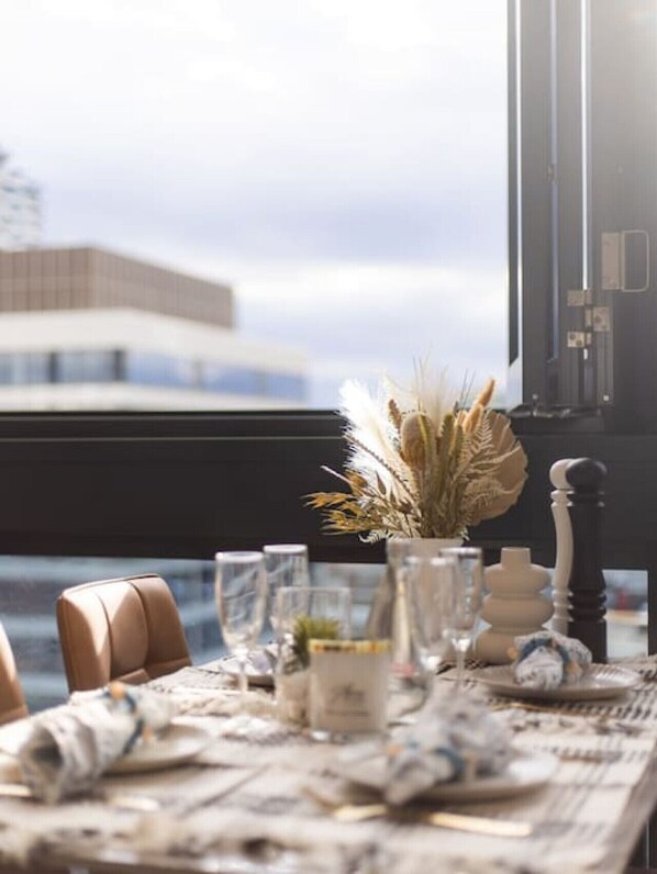 A dining table set with glasses, plates, and a vase with dried plants, overlooking a modern building.