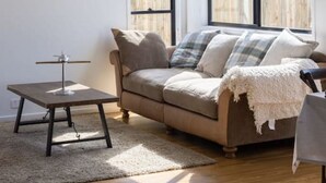 A living room with a beige sofa, a small table with a lamp, and a wall clock. There are large windows with a view of a wooden deck.