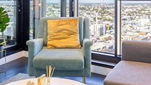 A modern living room with a large window offering a city view, a grey armchair with a yellow pillow, and a white coffee table with decorative items.
