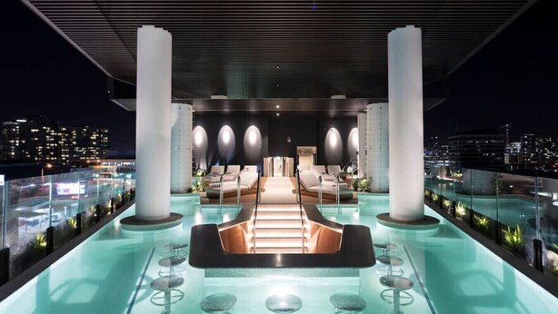 A rooftop lounge with a pool, seating areas, and a city view at night.