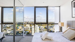 Une chambre d’hôtel moderne avec un grand lit, une table de chevet et une vue sur le paysage urbain grâce à de grandes fenêtres du sol au plafond.