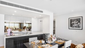 Une cuisine moderne avec un îlot central, une table à manger dressée avec des verres et des assiettes, et un coin salon confortable agrémenté d’un coussin moelleux pour chien.