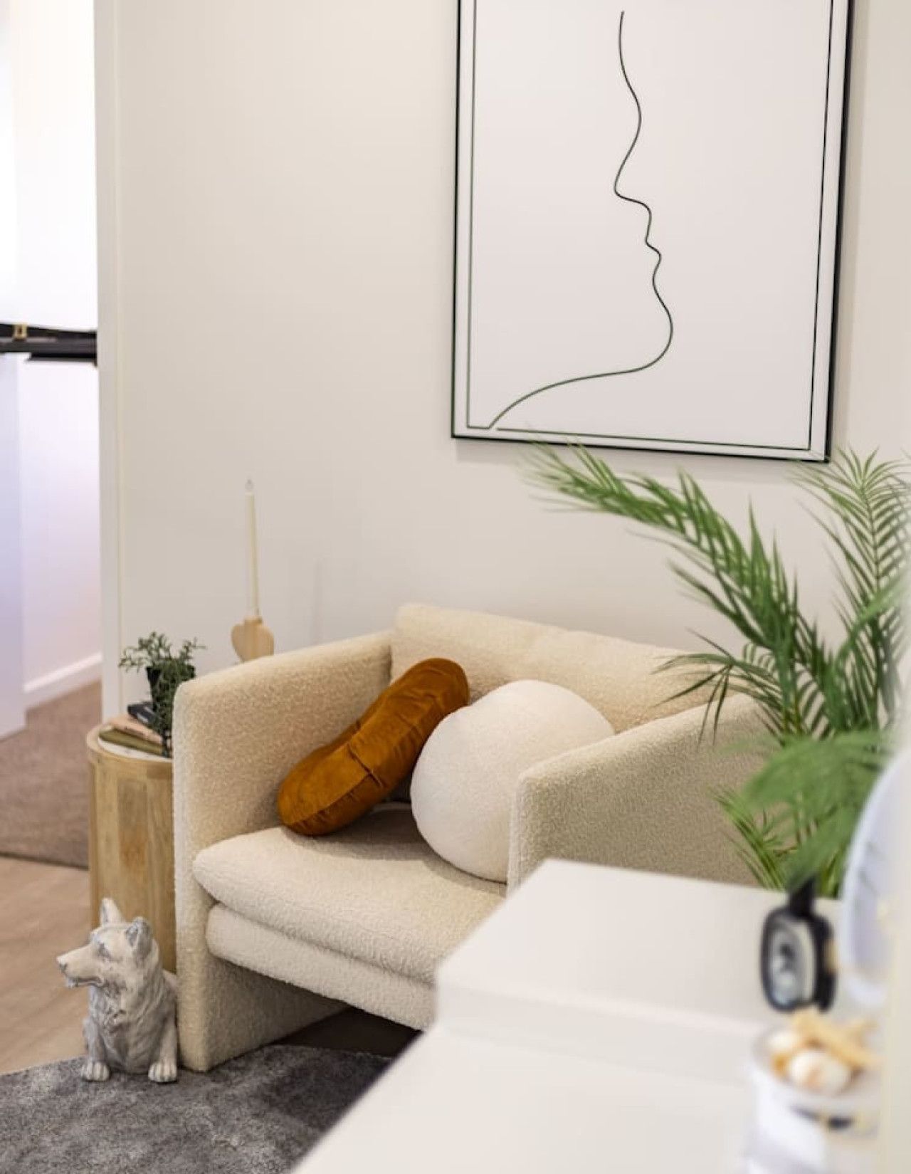 A living room with a sofa, a framed line drawing on the wall, a small wooden side table, a potted plant, and a dog statue.