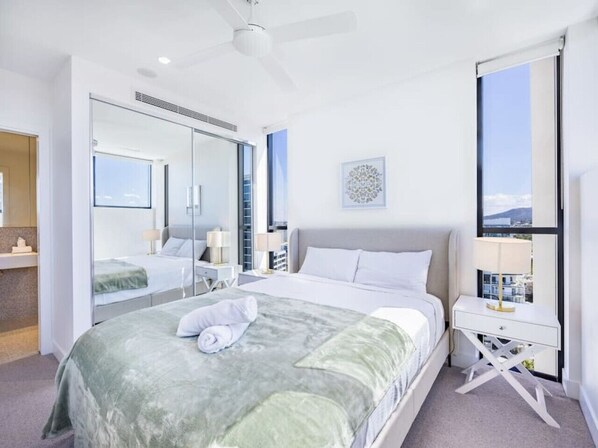 A modern hotel room with a large bed, a bedside table, a mirror, and a view of the city through a large window.