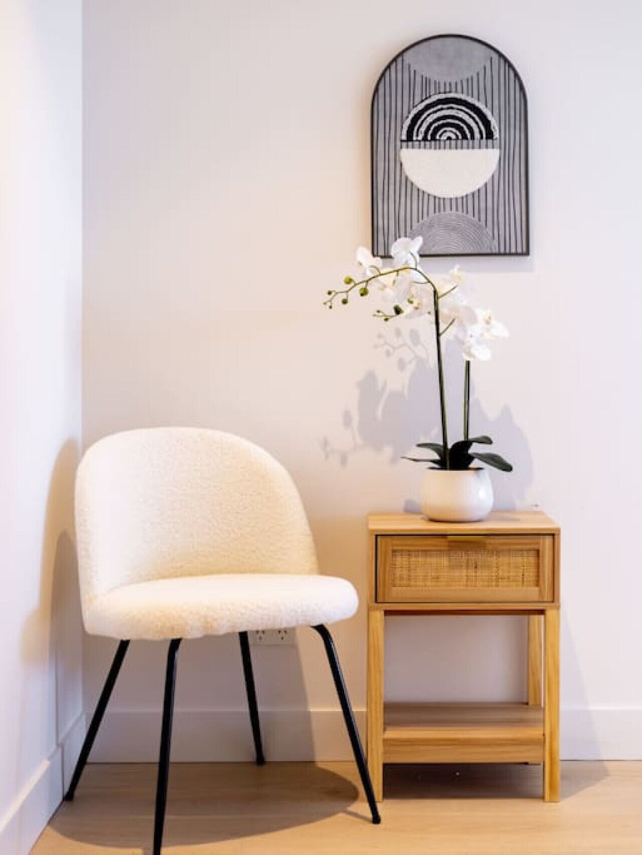 A white chair with black legs, a small wooden side table with a plant, and a wall-mounted abstract art piece.