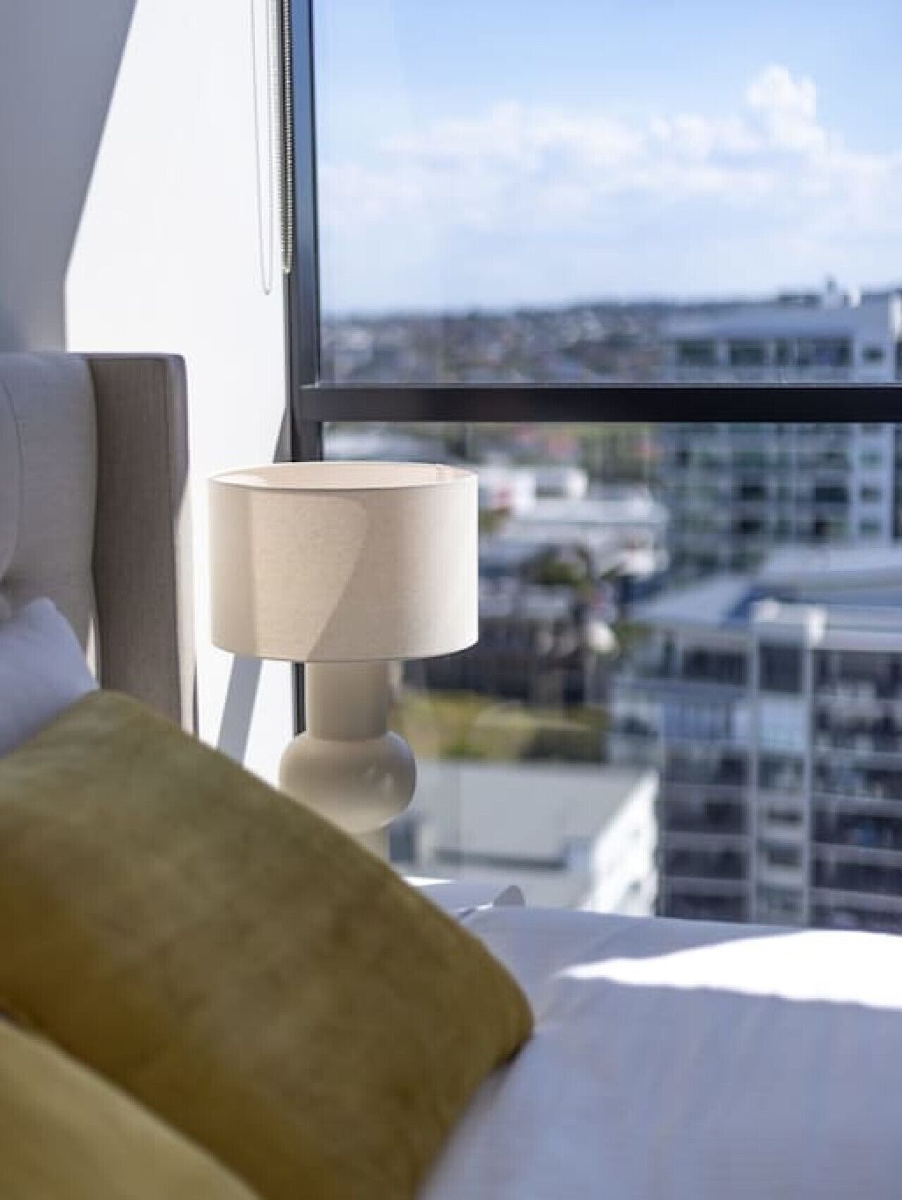 A hotel room with a bed, a lamp, and a view of a cityscape through a large window.