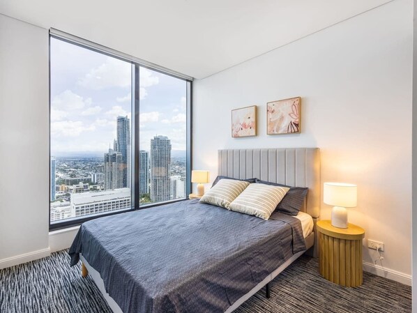 A modern hotel room with a large bed, two bedside lamps, a city view, and abstract wall art.