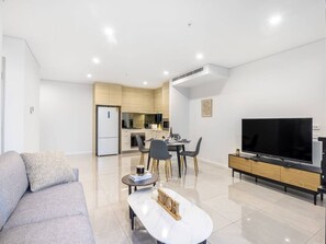 A modern living room with a sofa, a coffee table, a flat-screen TV, and a dining area with chairs and a table.