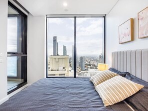 A bedroom with a large window, a bed with pillows, and a city view.
