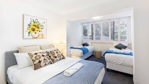 A hotel room with a large bed, two smaller beds, a window with blinds, a ceiling light, and a wall-mounted artwork.