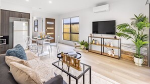 A modern living room with a sofa, coffee table, and a TV on a stand. There is a dining area with chairs and a table, and a kitchen area with a refrigerator and microwave.