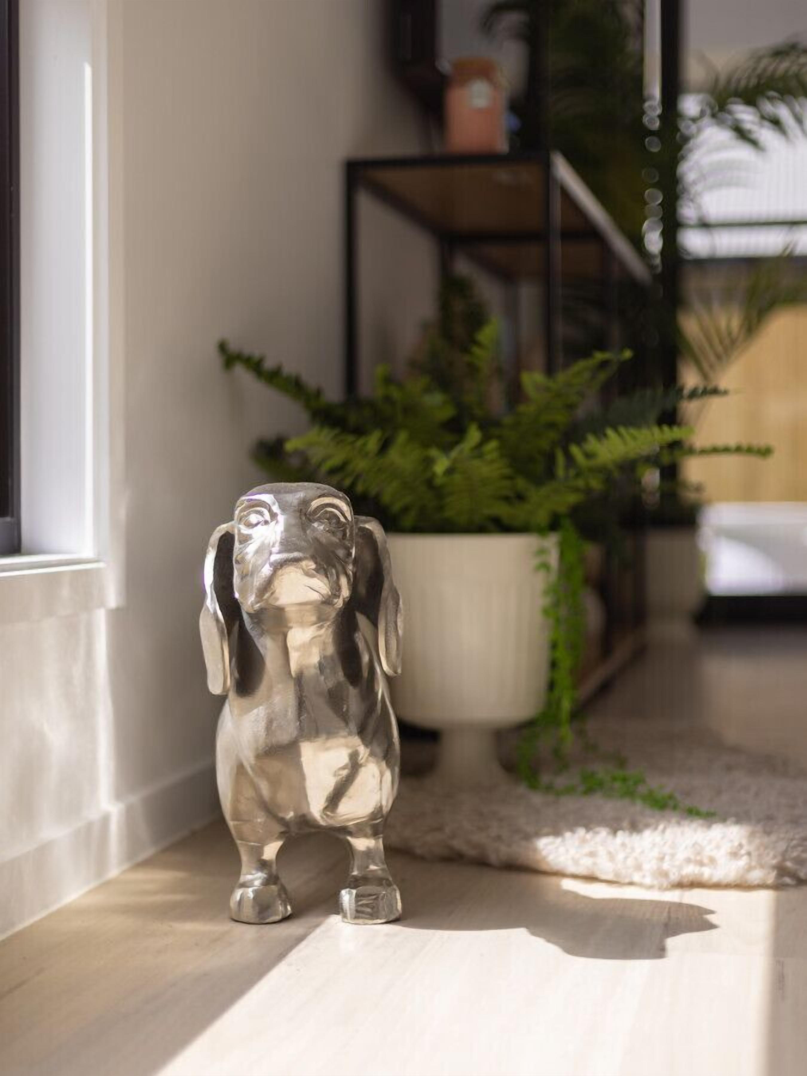 Una figurita de perro plateado sobre un piso de madera, cerca de una ventana, con una planta en maceta y un estante negro al fondo.