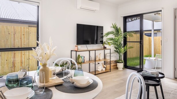 A modern living room with a dining area, a TV mounted on the wall, and a sliding glass door leading to a garden.