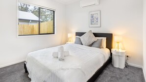 A neatly arranged bedroom with a bed, two bedside lamps, a ceiling fan, and a window with a view of a fence and trees.