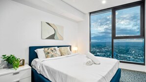 A modern hotel room with a large bed, a nightstand, a lamp, a view of the city through large windows, and a wall-mounted abstract art piece. - Cloudview Retreat | Luxe Skyhome Experience (Surfers Paradise)