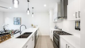 A modern kitchen with white cabinetry, a central island with a sink, stainless steel appliances, pendant lighting, and a framed artwork on the wall. - 4BR Cozy house 5mins to beach (Surfers Paradise)