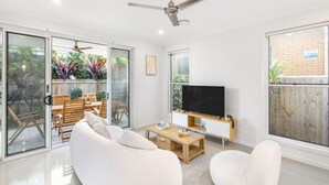 A modern living room with a flat-screen TV, white sofas, a wooden coffee table, and a ceiling fan. - 4BR Cozy house 5mins to beach (Surfers Paradise)