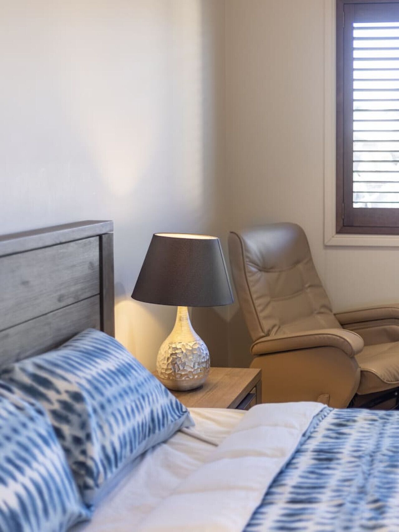 Un lit bien fait, agrémenté d’oreillers à motifs, une lampe de chevet et un fauteuil en cuir dans une pièce dotée d’une fenêtre.