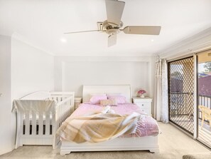 Ein Schlafzimmer mit Bett, Babybett, Nachttisch, Deckenventilator und einer Schiebeglastür nach draußen.