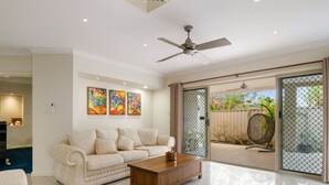 A spacious living room with a white sofa, a wooden coffee table, and a ceiling fan. There are three colorful paintings on the wall and a sliding glass door leading to an outdoor area with a wicker chair. - Private Cinema! 5Beds Spacious Villa Near Shops (Calamvale)