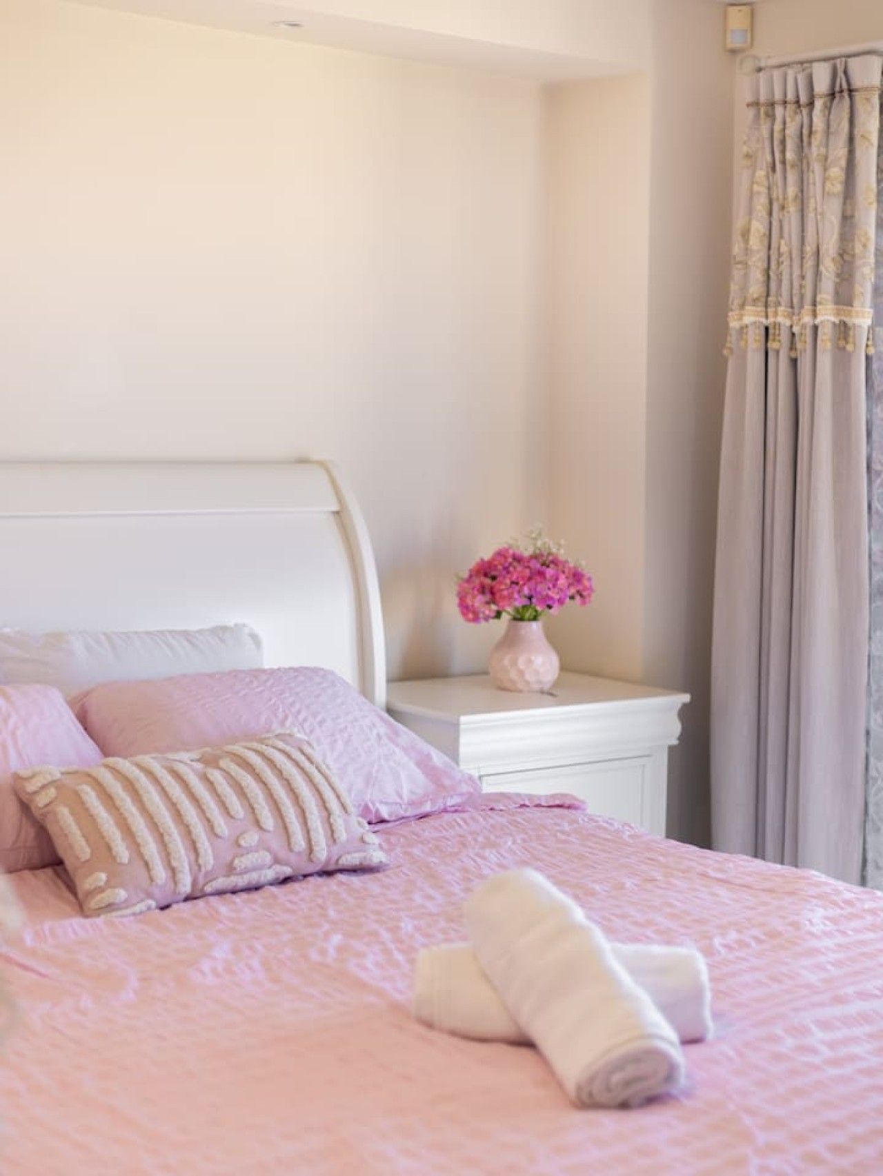 Un lit avec une literie rose, une tête de lit blanche et une table de chevet ornée d’un vase de fleurs.