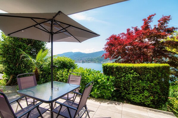 An outdoor patio with a table and chairs, a large umbrella, and a view of a lake and mountains. - Studio apartment with lakeview terrace (LA PALMA 3) (Collina d'Oro)