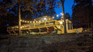 Uma cabana de madeira com vários níveis, terraço coberto e área de estar externa, cercada por árvores e uma parede de pedra.