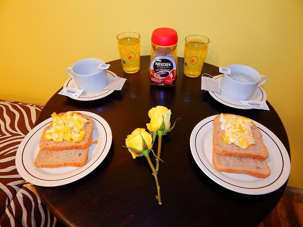 Une table avec deux assiettes de toasts garnies d’œufs brouillés, une bouteille de Nescafe, deux tasses de thé et un bouquet de roses jaunes.