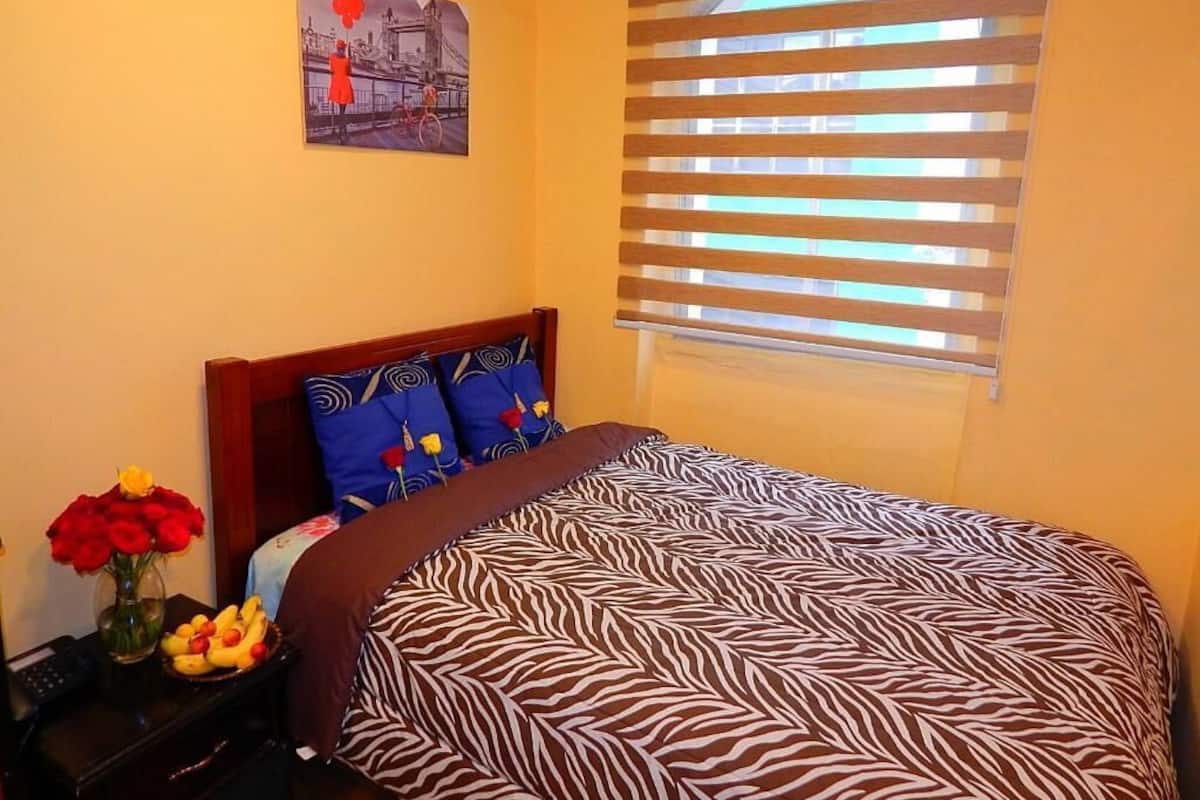 A bedroom with a zebra-patterned bedspread, a wooden headboard, a nightstand with a vase of red roses and a bowl of fruit, and a window with horizontal blinds.