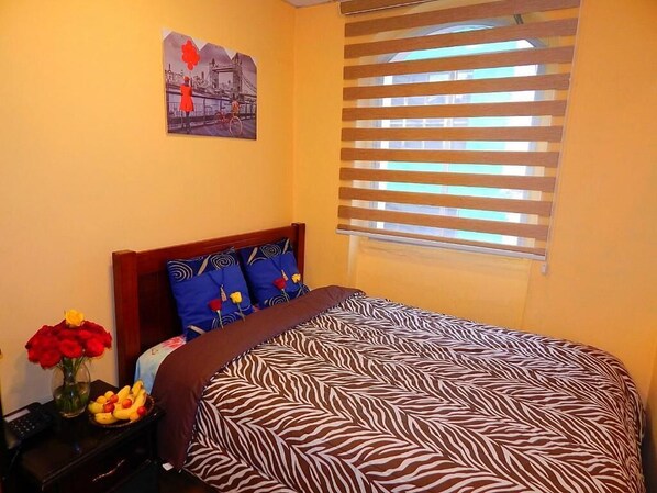 A bedroom with a zebra-patterned bedspread, a wooden headboard, a nightstand with a vase of red roses and a bowl of fruit, and a window with horizontal blinds.