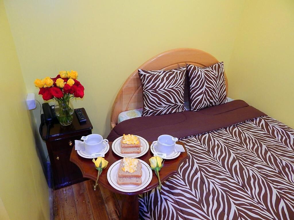 Una cama bien hecha con una colcha estilo cebra, una mesita de noche con un florero y dos platos con rebanadas de pastel y tazas de té.
