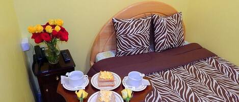 Una cama bien hecha con una colcha estilo cebra, una mesita de noche con un florero y dos platos con rebanadas de pastel y tazas de té.