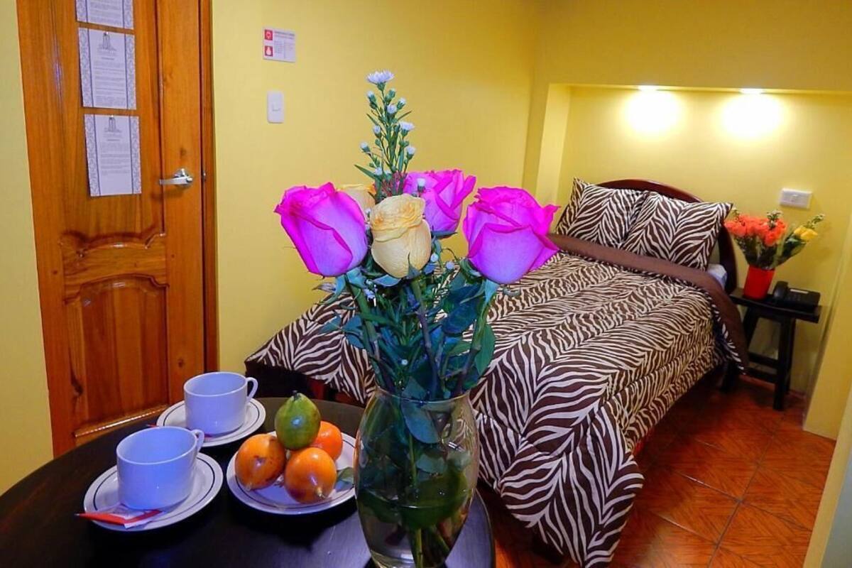 A room with a zebra-patterned bed, a wooden door, a table with cups and fruit, and a vase with flowers.