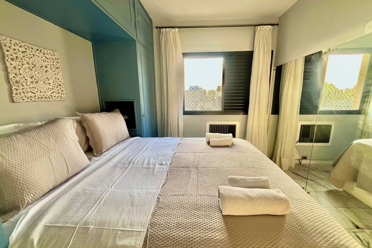 A neatly made bed with a white comforter and pillows, a decorative wall piece, and a window with curtains.