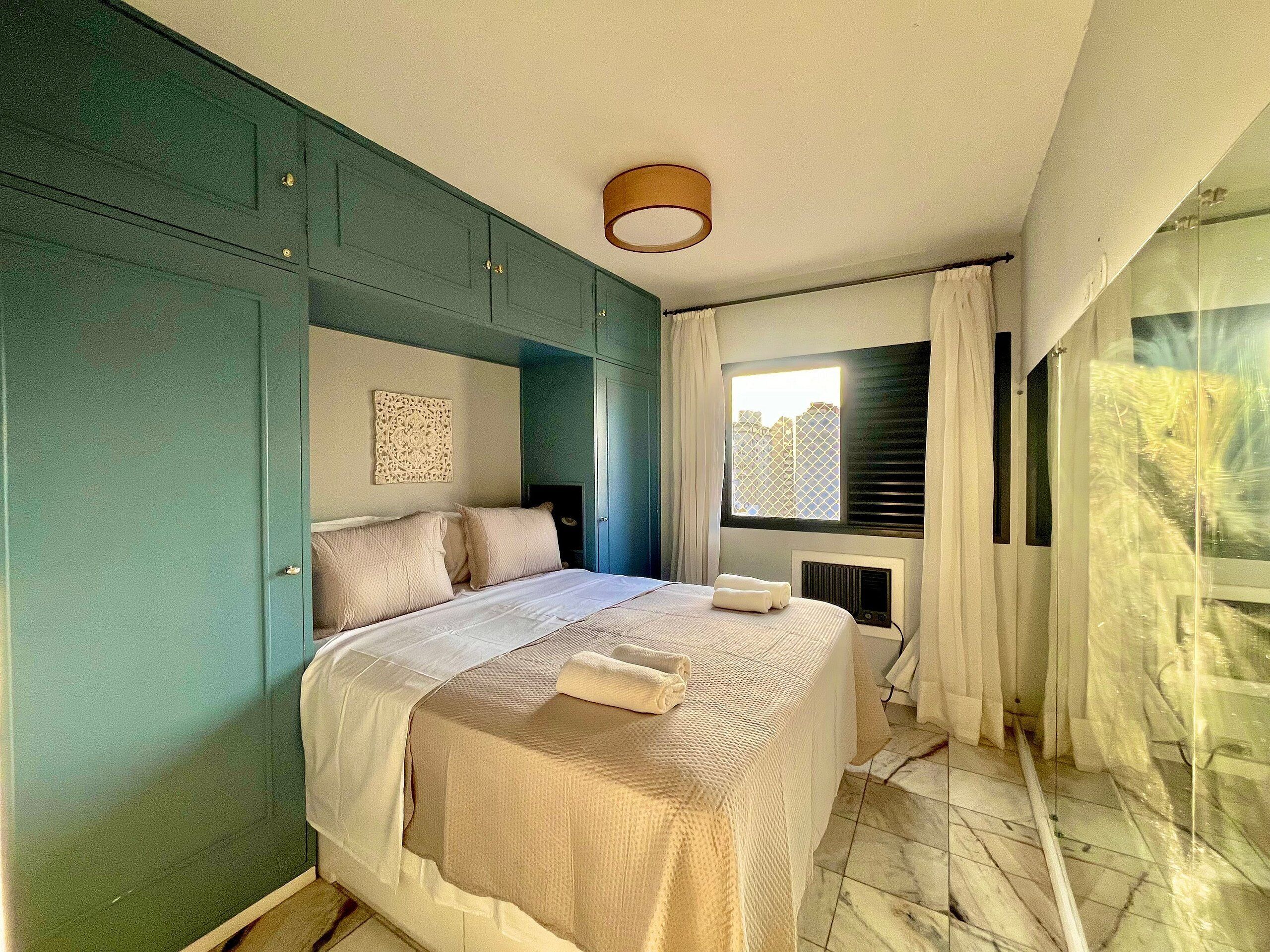 A bedroom with a bed, a radiator, a window with blinds, and a view of buildings outside.