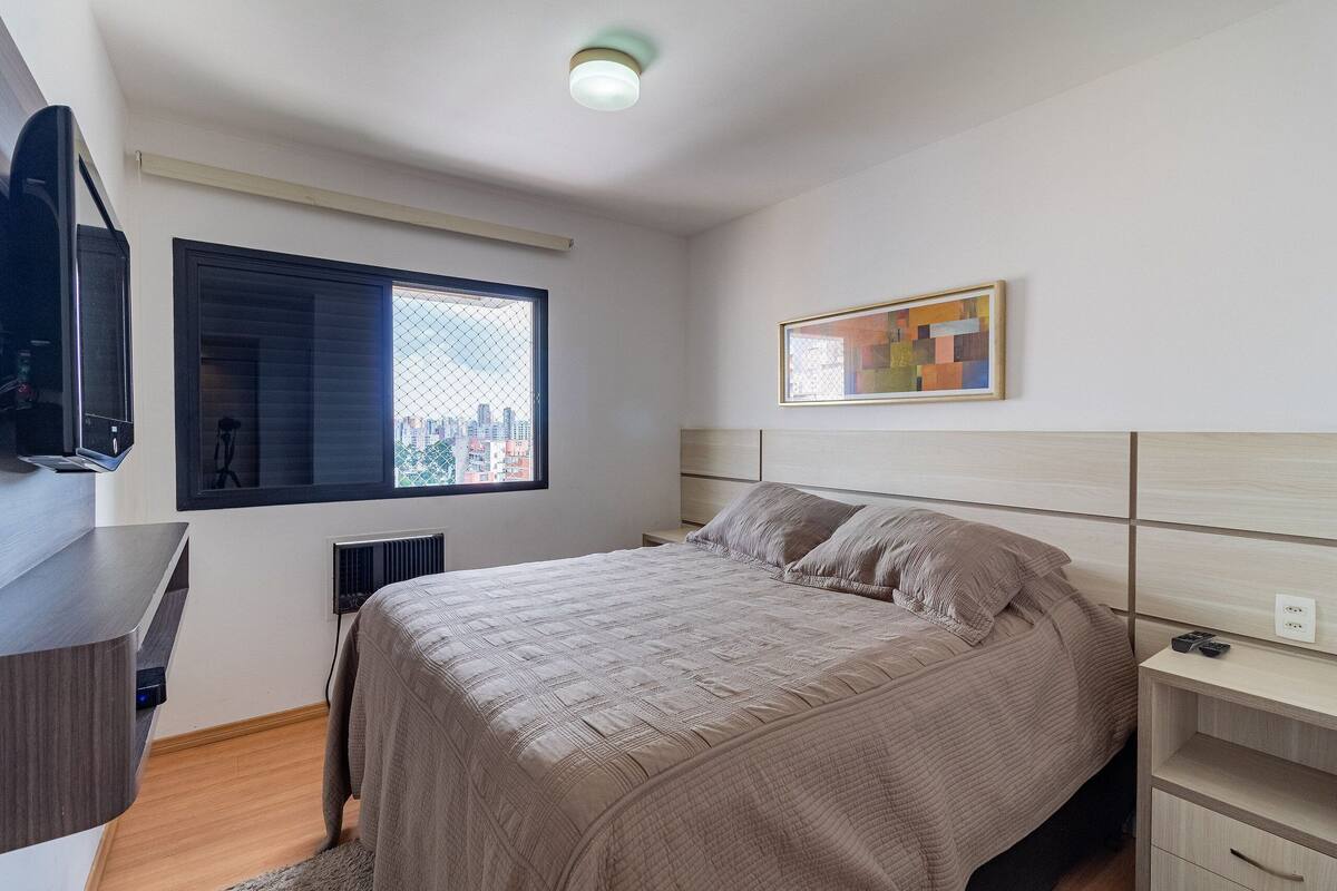 A modern bedroom with a bed, a TV, a window with a city view, and a framed artwork on the wall.