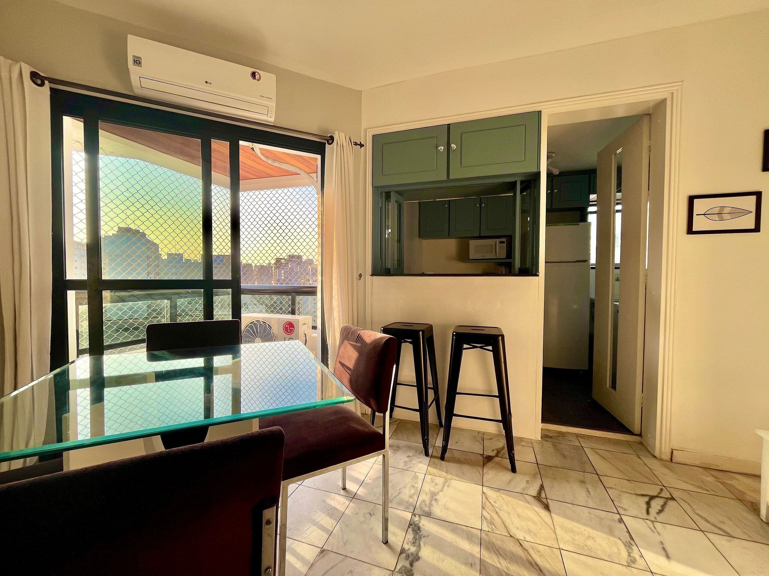 A dining area with a glass table, two bar stools, a cushioned chair, and a view through a window with a curtain.
