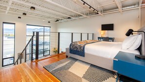 A modern hotel room with a large bed, a desk, and a view of the mountains through the windows. - The Lofty View:Midtown Bozeman (Bozeman)
