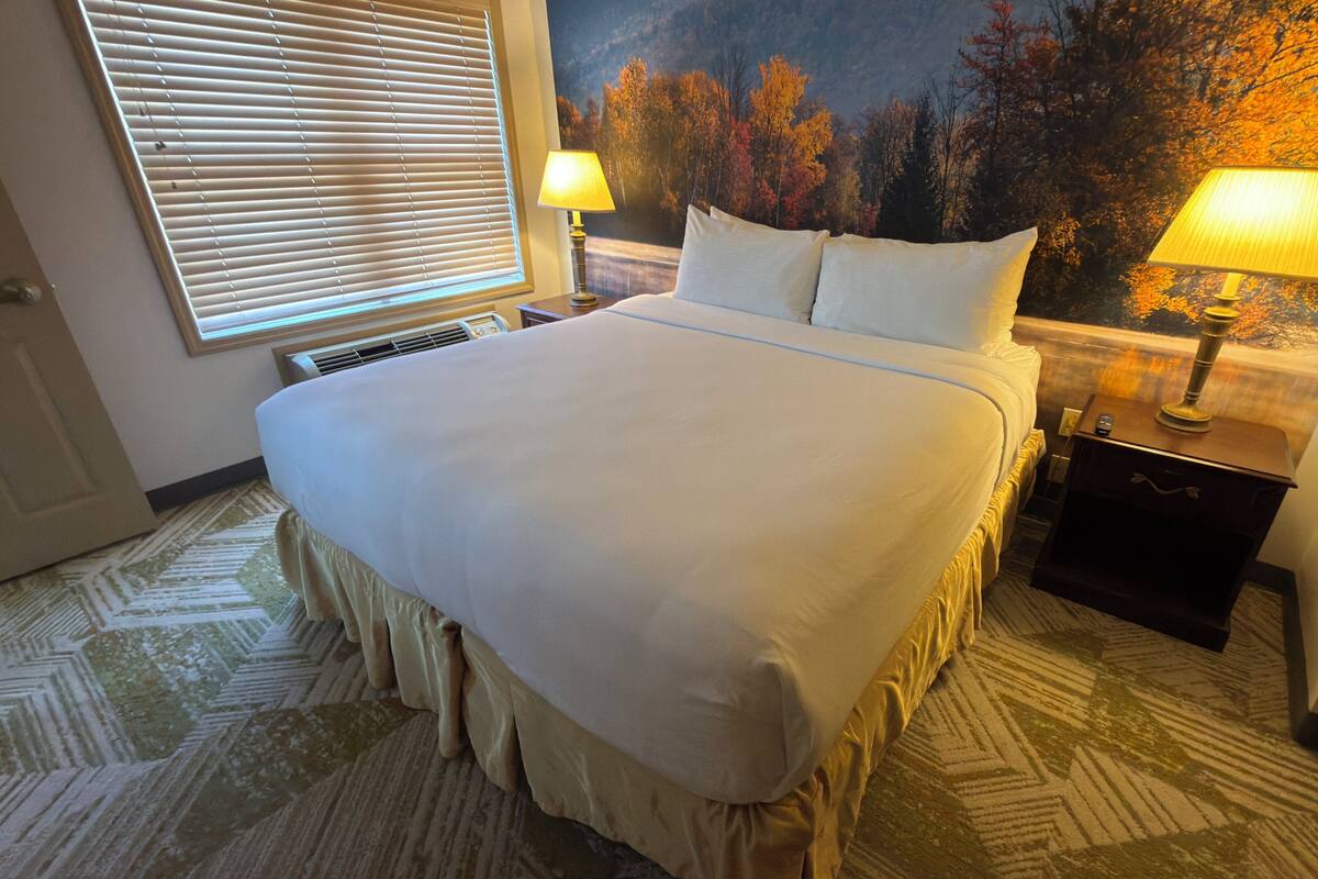 Ein Hotelzimmer mit einem großen Bett, zwei Nachttischlampen, einem Nachttisch, einem Fenster mit Jalousien und einem Wandgemälde.