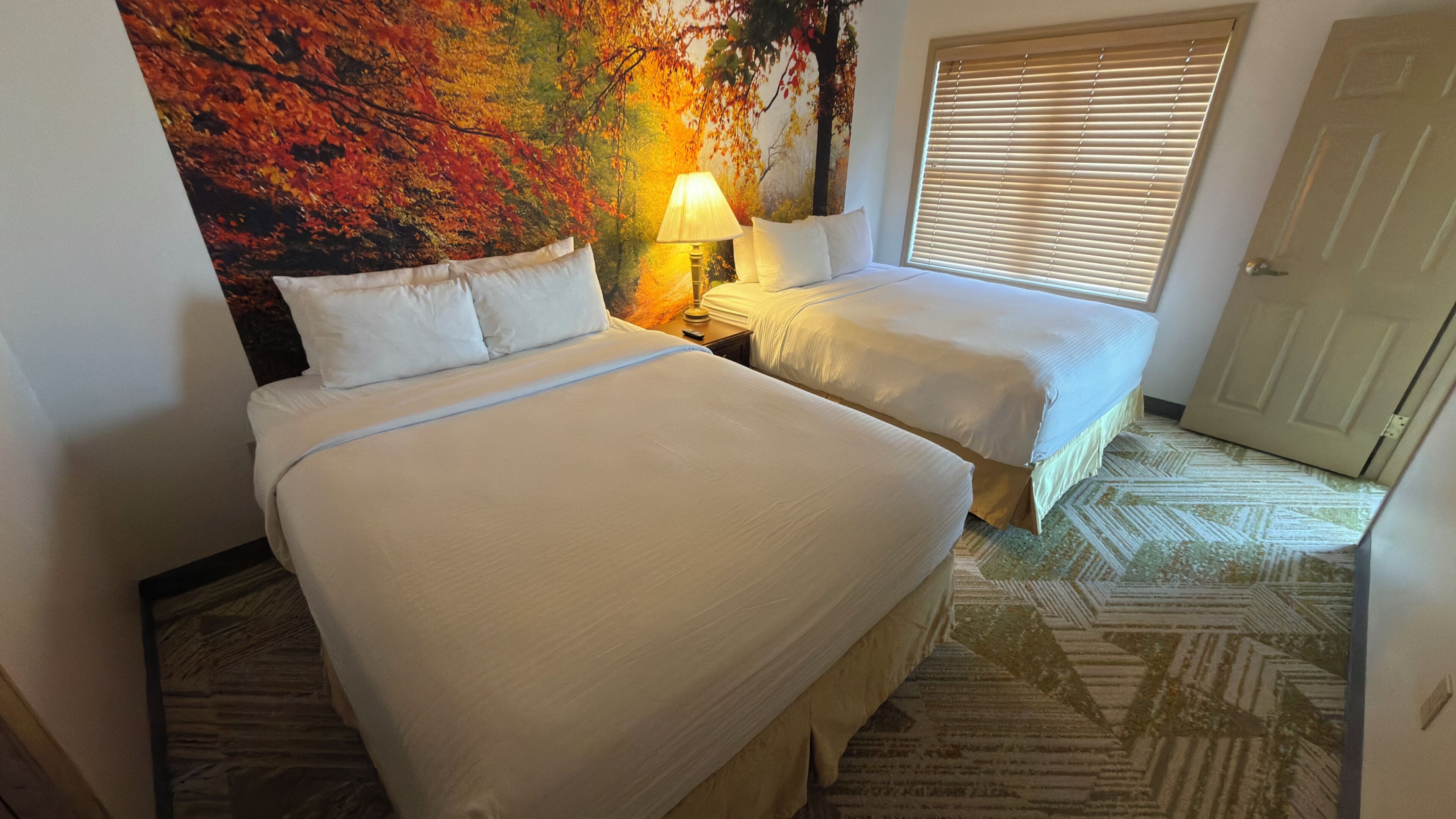 Habitación de hotel con dos camas, una mesita de noche con lámpara, un mural grande con motivos otoñales y una alfombra estampada.