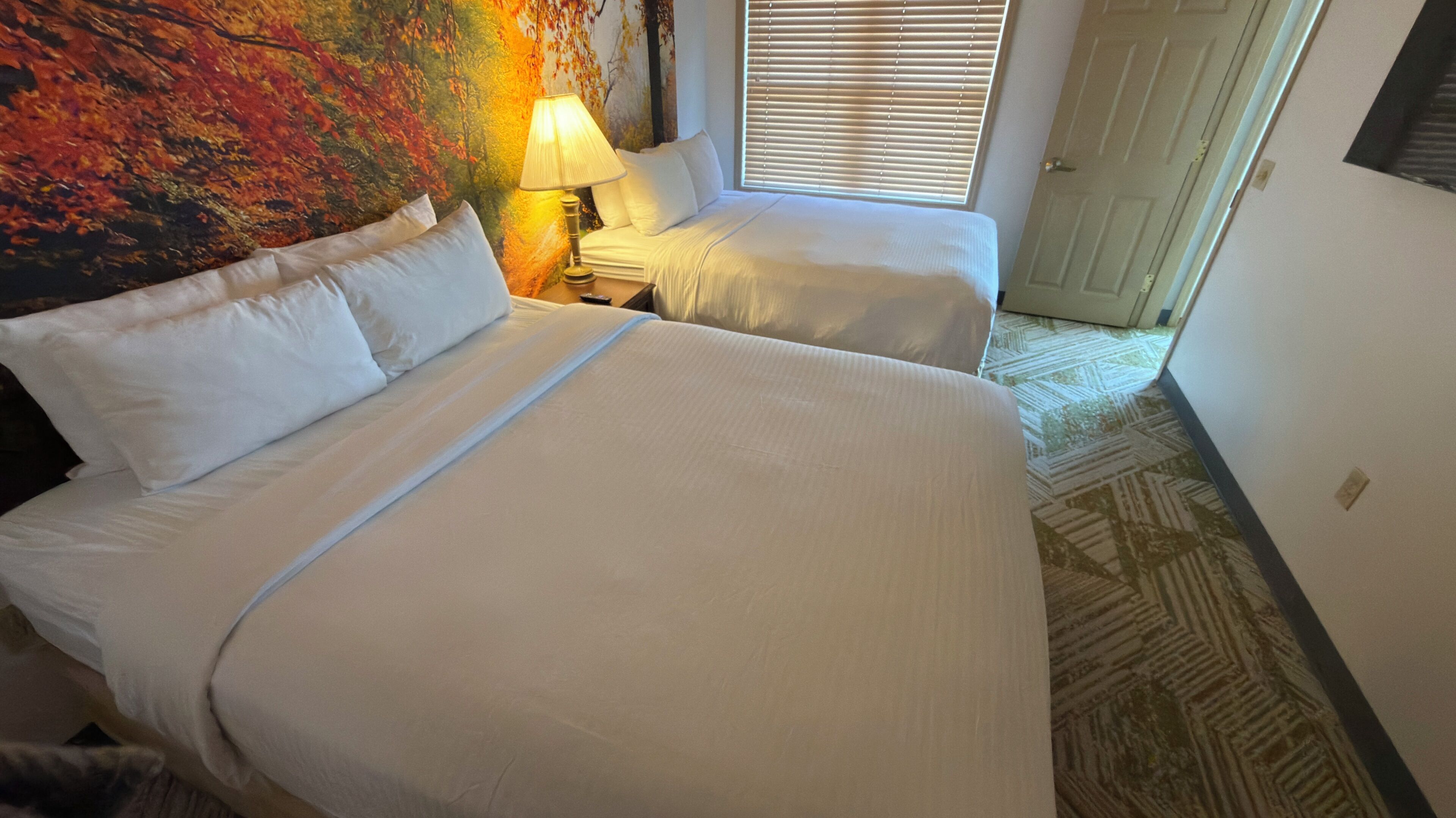 Habitación de hotel con dos camas, un cuadro en la pared, una lámpara y una ventana con persianas.