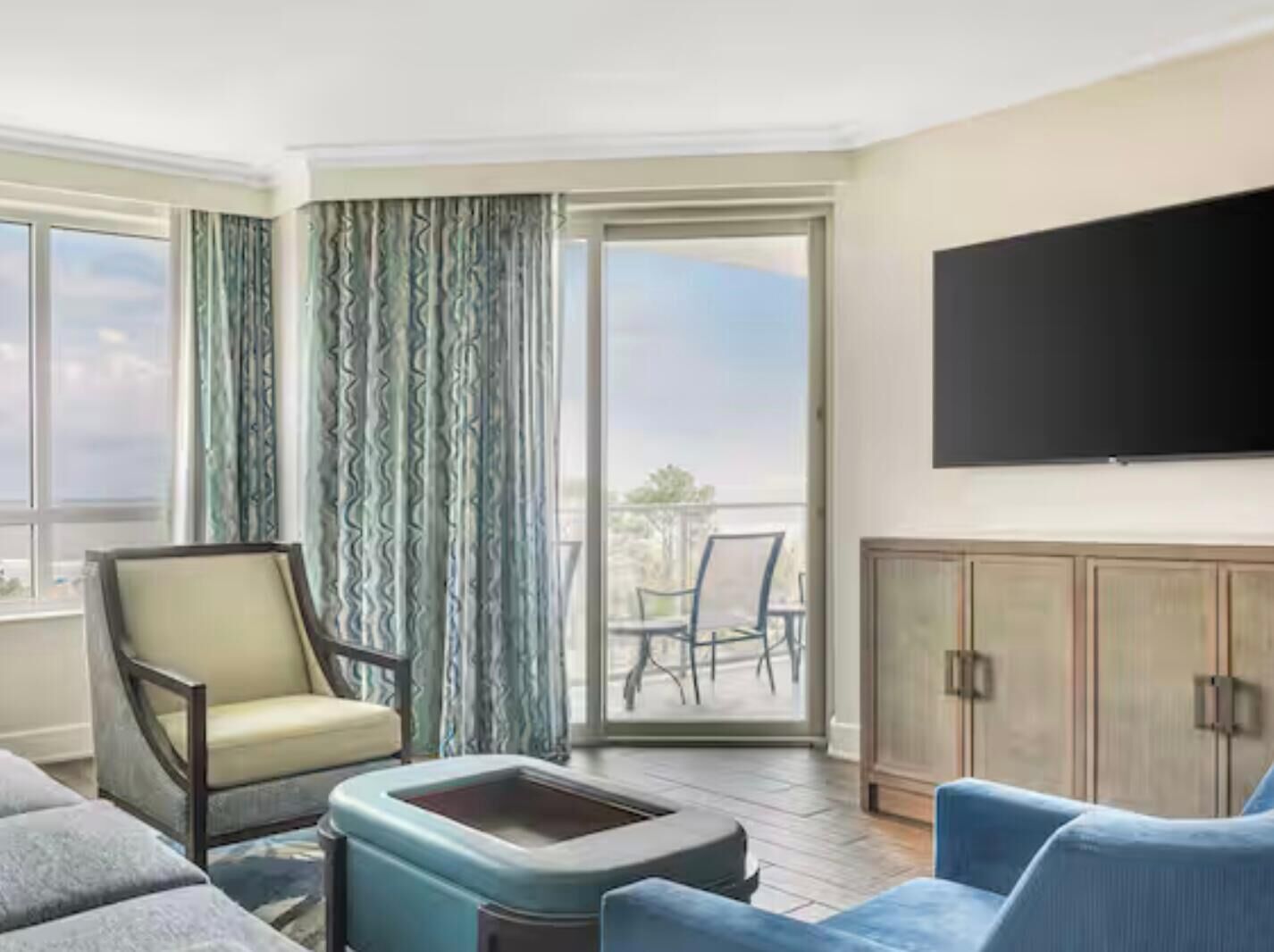Une chambre d’hôtel comprenant un canapé, un fauteuil, un téléviseur à écran plat fixé au mur, une porte-fenêtre coulissante donnant sur un balcon avec des chaises, et un meuble en bois.