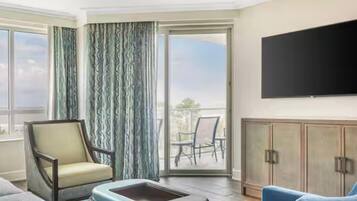 Une chambre d’hôtel comprenant un canapé, un fauteuil, un téléviseur à écran plat fixé au mur, une porte-fenêtre coulissante donnant sur un balcon avec des chaises, et un meuble en bois.